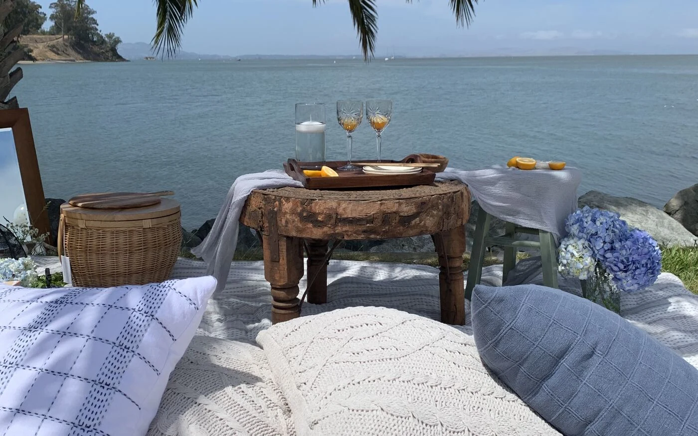 Waterfront picnic setup with a carved wood table, hydrangeas and knit pillows